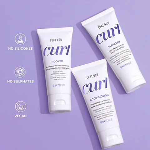Three white tubes of Curl Wow hair products are displayed on a light purple background, with the visible text including CURL WOW curl HOOKED Shampoo with Root-Lock Tech Shampooing fixation des racines Residue-free, sulfate-free formula Formule sans résidus, sans sulfates 60 ml e 2.0 FL OZ, CURL WOW curl FLO-ETRY Vital Natural Serum Sérum naturel vital Delivers critical moisture levels. Clinically proven, 84% less breakage. Apporte une hydratation vitale. Testé cliniquement, 84% moins de casse. 60 ml e 2.0 FL OZ, CURL WOW curl COCO-MOTION Lubricating Conditioner Après-shampooing lubrifiant Restores fluidity + elasticity for super-spring curls. Rend vos cheveux plus fluides et plus élastiques pour des boucles rebondies. 60 ml e 2.0 FL OZ, and the feature icons reading NO SILICONES, NO SULPHATES, VEGAN.